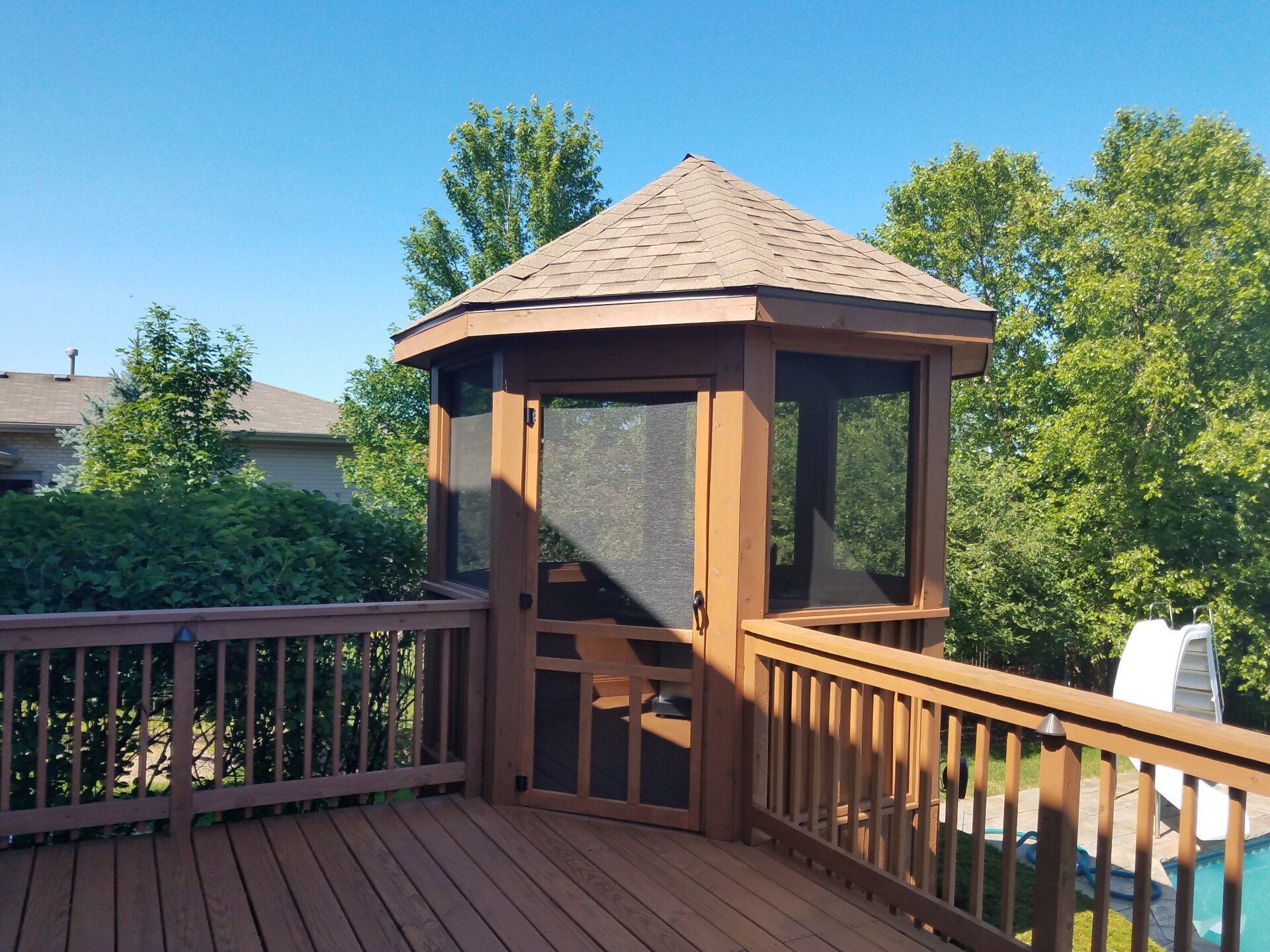 Wooden gazebo on a deck with brown roof, screening, and surrounding trees under a blue sky.