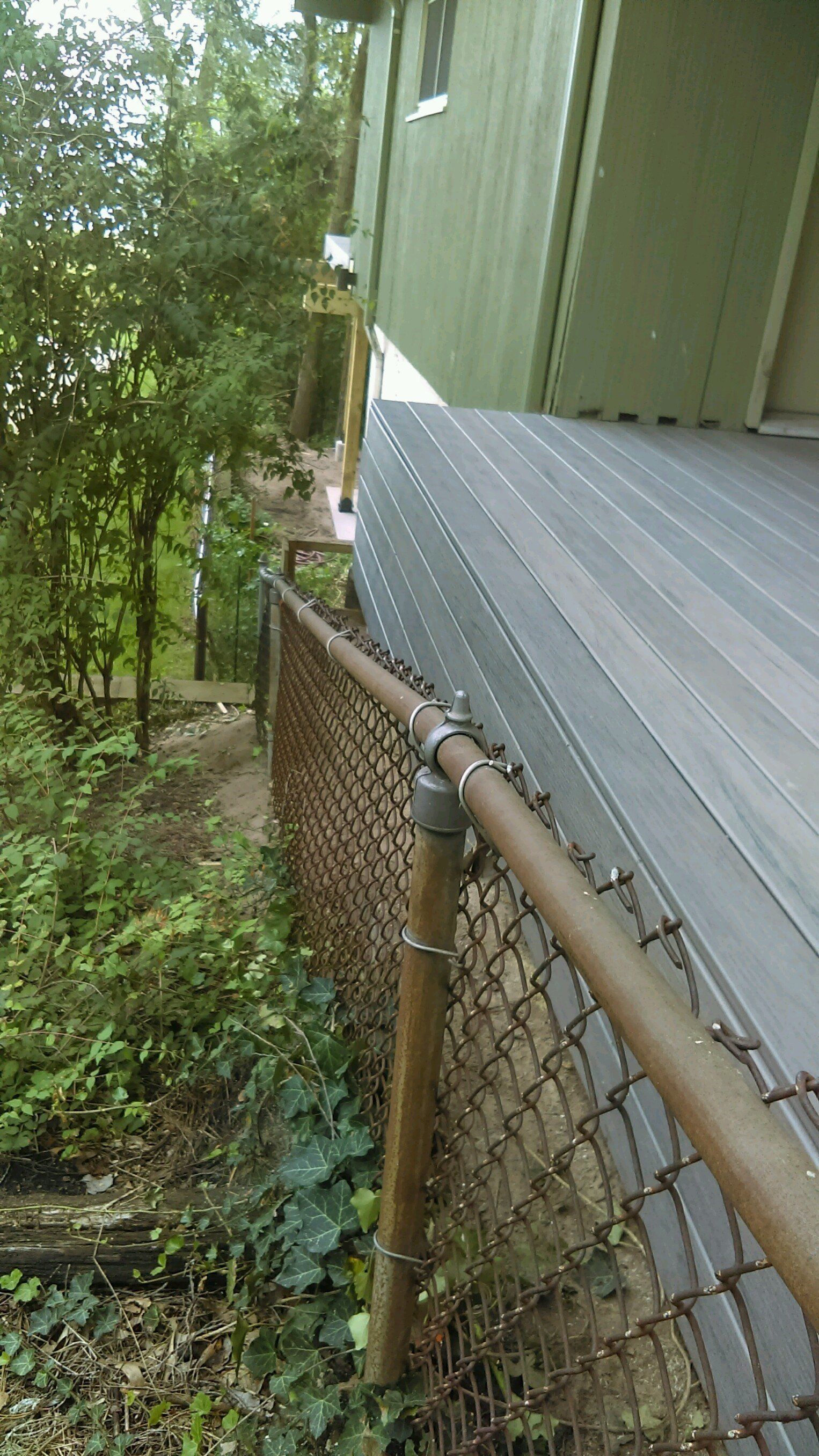 Chain-link fence borders a wooden deck next to a green building, a pathway slopes down into trees.