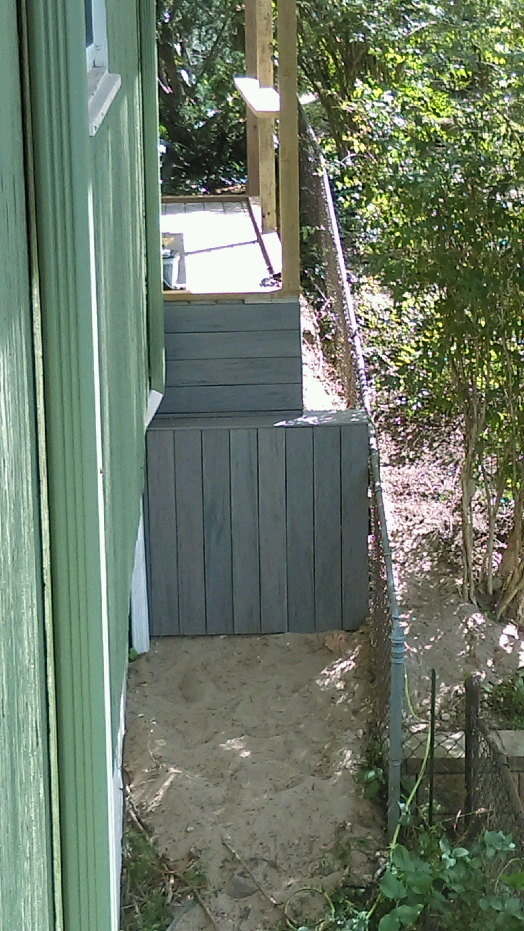 Wooden steps leading up to a deck, set against a green house, with a sandy area below.