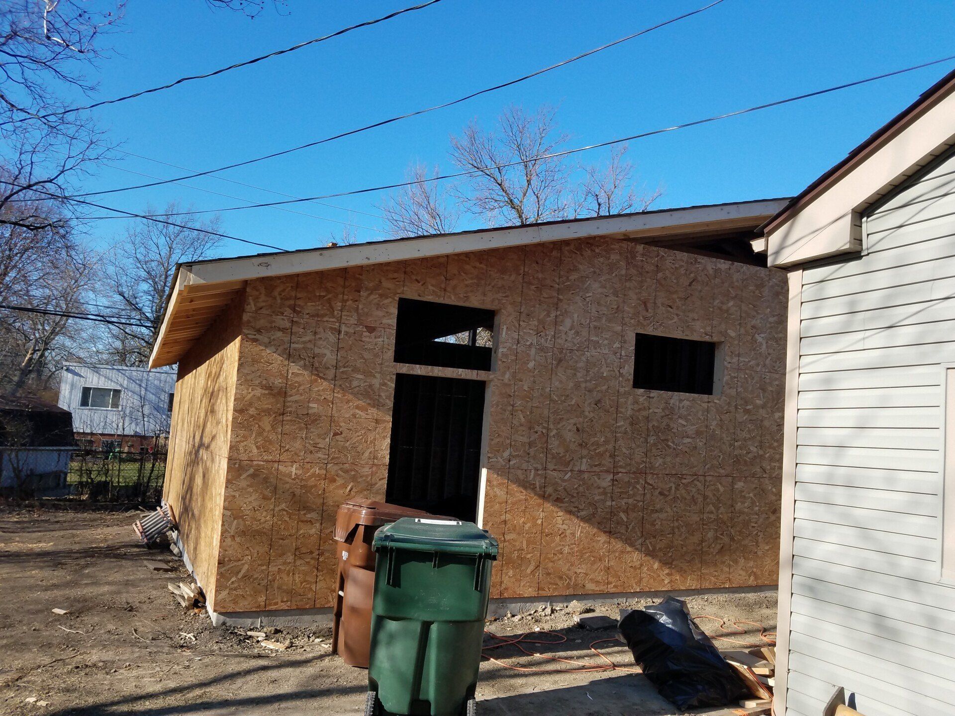 Exterior view of a building under construction, brown siding, a green trash bin, and blue sky.