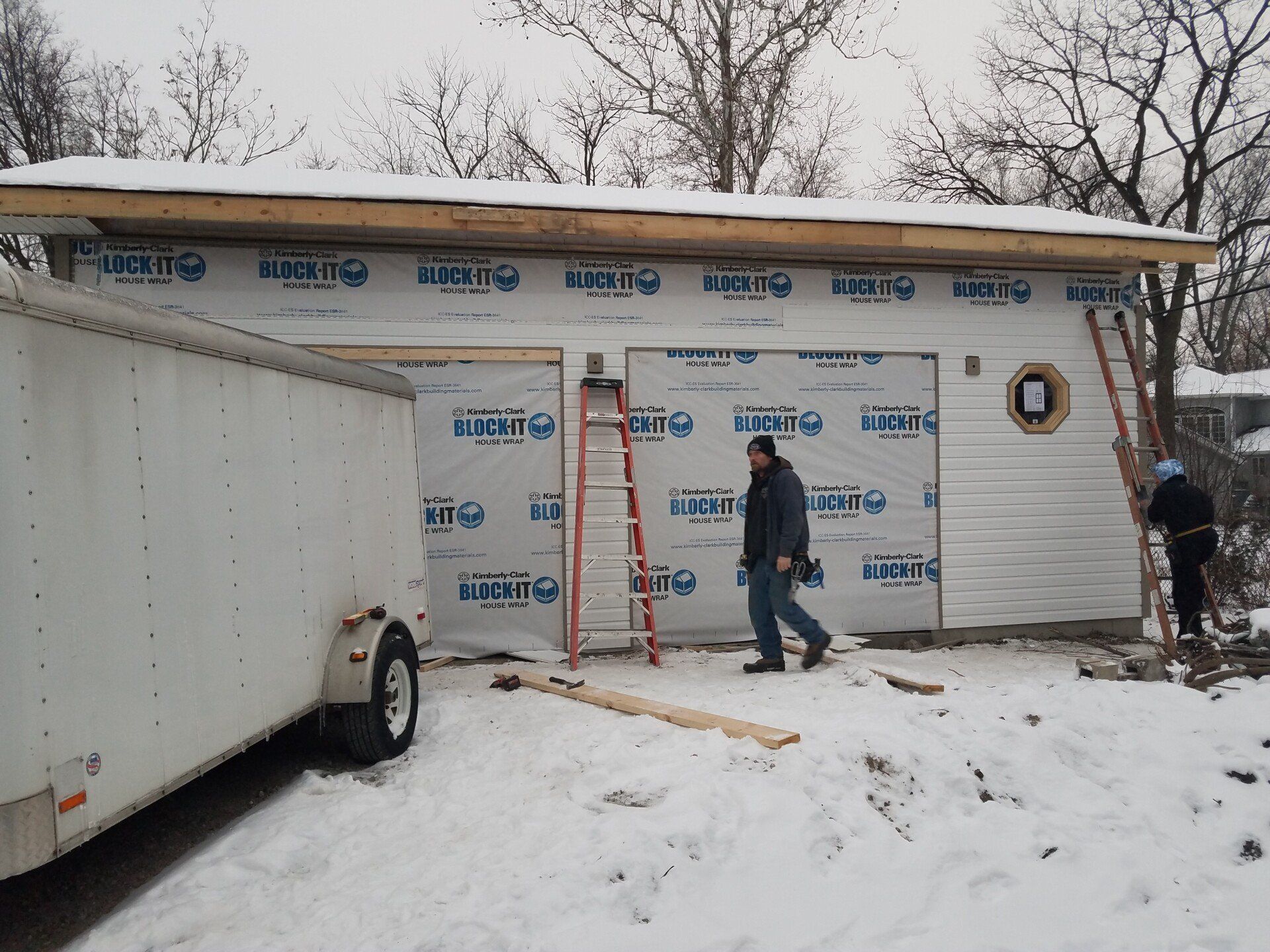 Garage under construction, two people working, white trailer, snow on the ground.