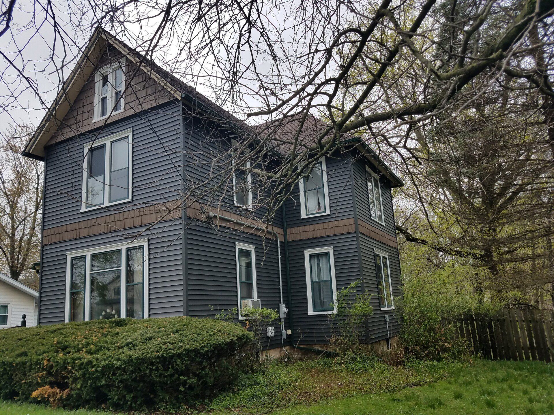 Two-story house with dark blue siding, brown trim, and a small front yard with overgrown bushes.