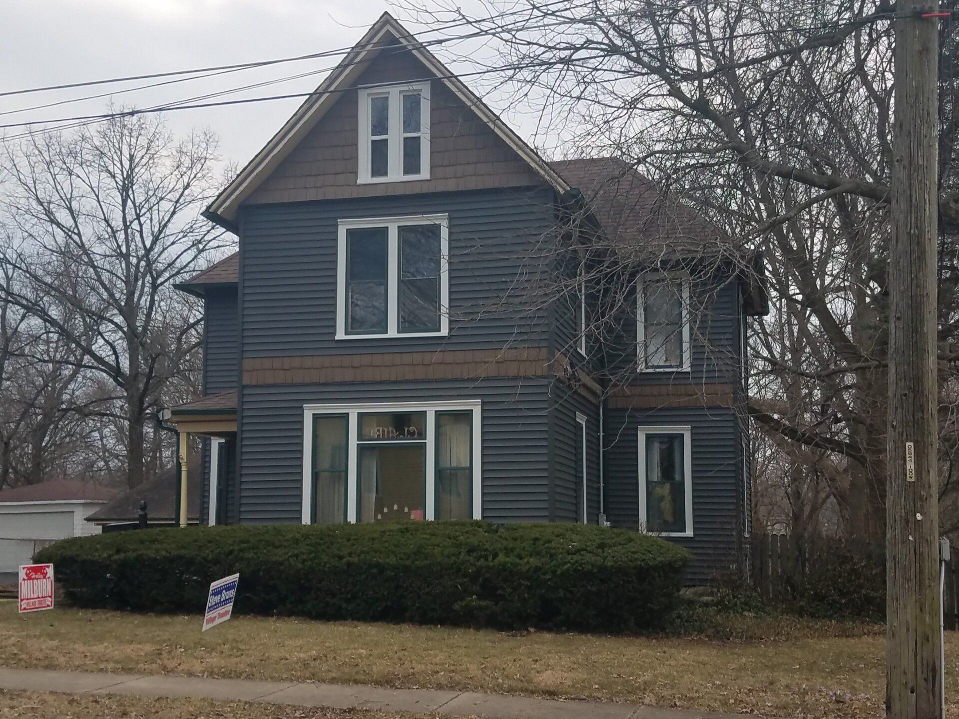 Two-story house with dark gray siding, brown trim, and a small front yard with bushes.