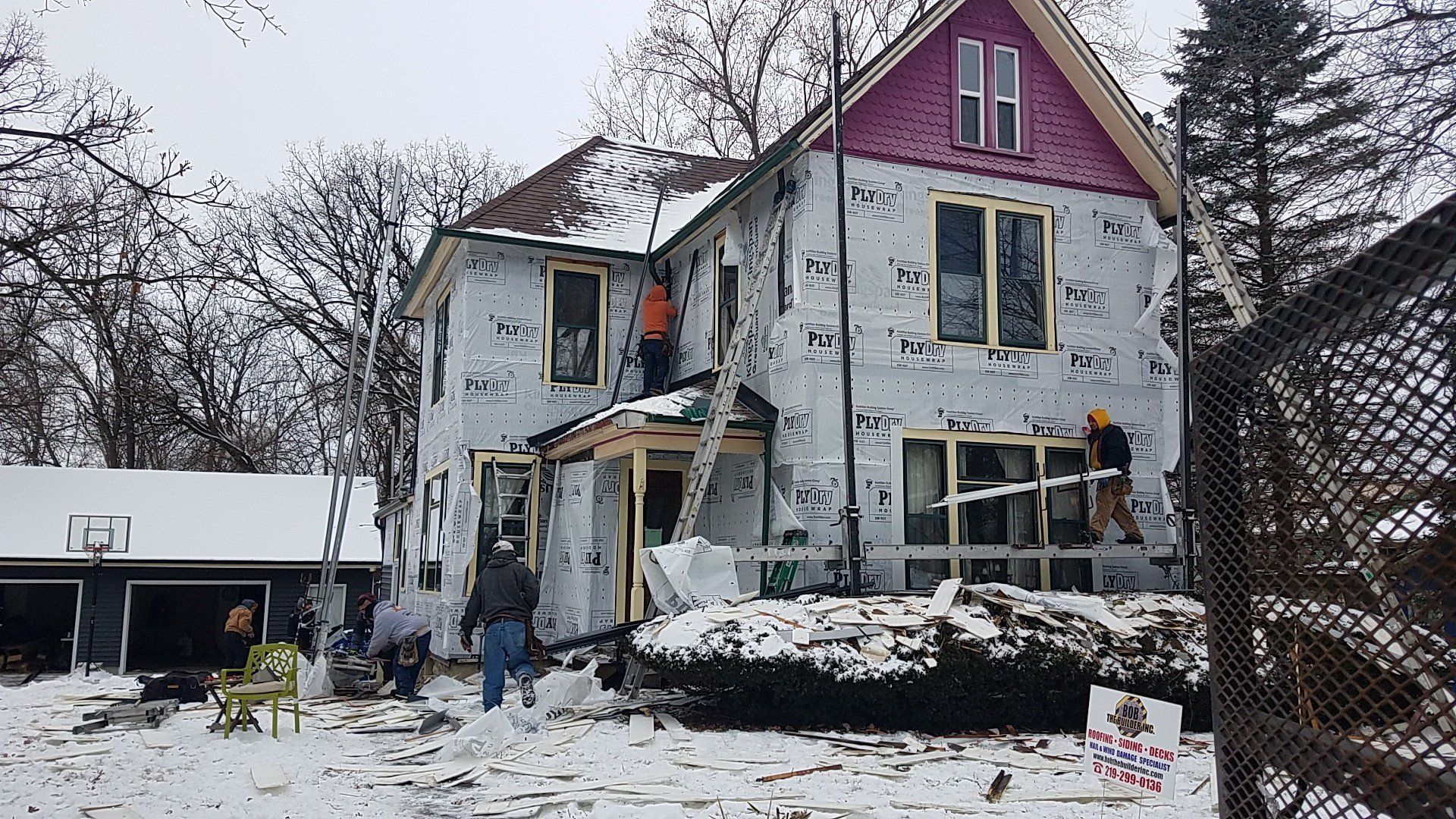 House under construction, covered in wrap; workers outside in snowy weather.