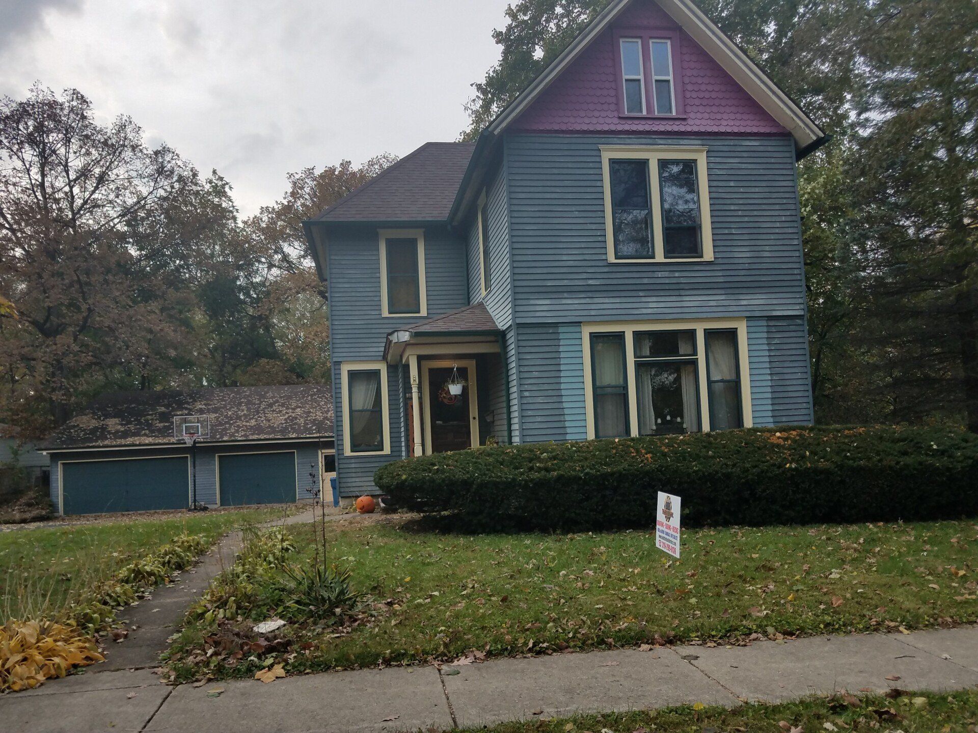 Blue two-story house with detached garage; pink and brown trim, overcast sky, lawn with bushes.