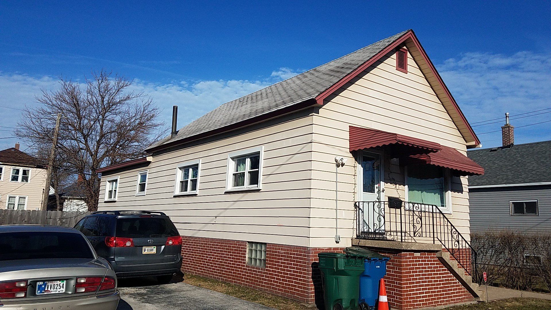 House with tan siding, red brick foundation, awning over door, cars parked in front.