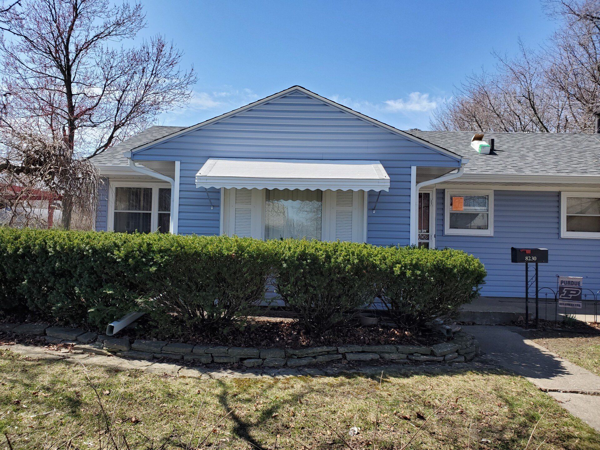 Blue house with white awning, hedge, and a small yard.