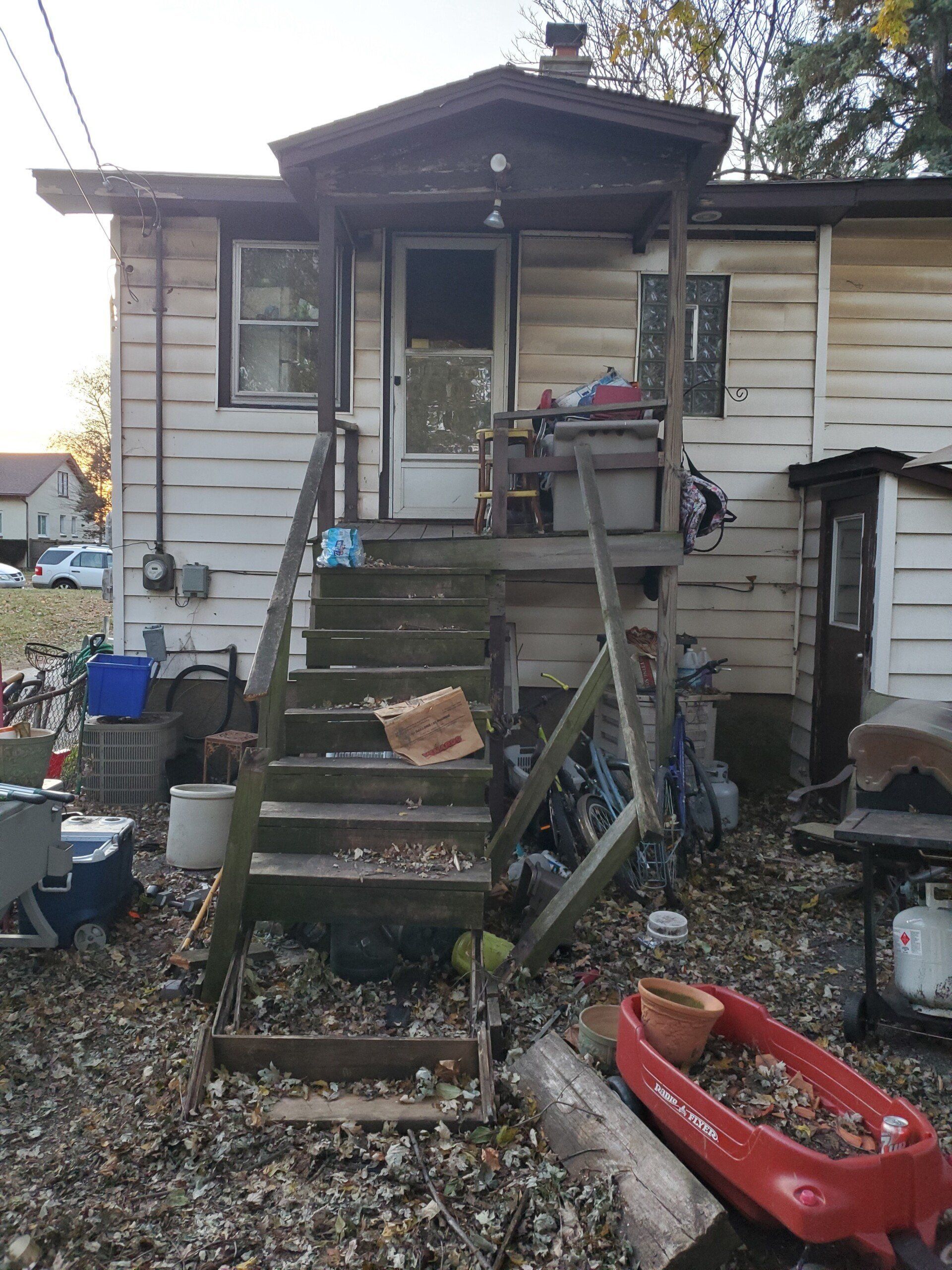 Dilapidated wooden steps lead to a porch door with clutter and debris scattered around.