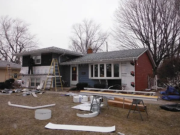 House undergoing siding replacement; exposed sheathing, materials and ladder on lawn.