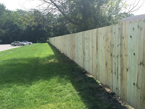 Wooden fence along a grassy area, with trees and cars visible in the background on a sunny day.