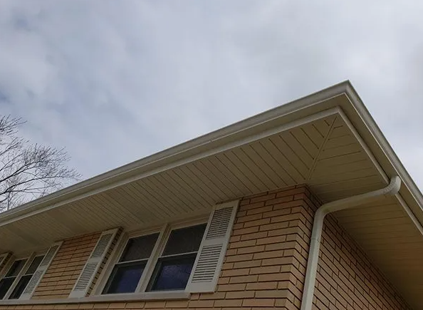 Beige brick house corner with white trim, gutters, and shutters. Overcast sky.