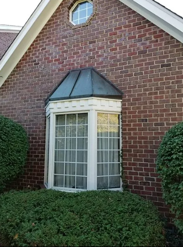 Brick building with bay window, green bushes below. A small, round window above the bay window.