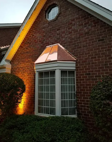 Copper-roofed bay window on a brick building, lit with warm exterior lighting.