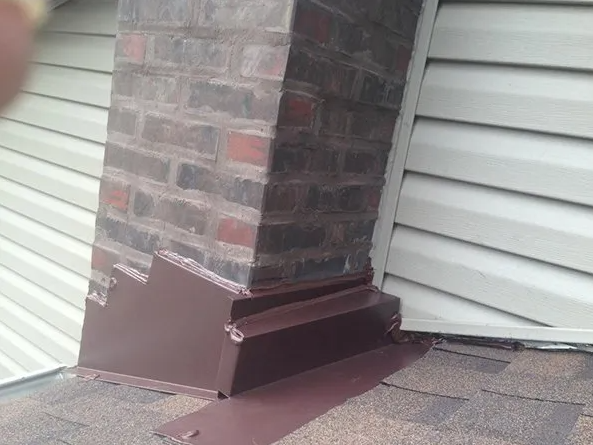 Brick chimney with brown metal flashing on a shingled roof, next to beige siding.