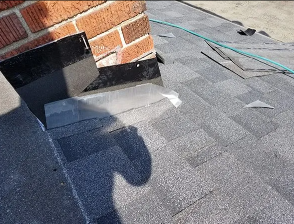 Close-up of a roof with grey shingles, brick chimney, and metal flashing where the roof meets the chimney.