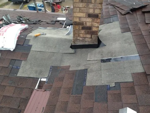 Roof with exposed underlayment around a brick chimney, shingles partially removed.
