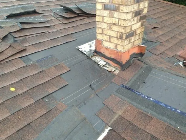 Damaged asphalt shingle roof with a brick chimney. Flashing is visible, shingles are missing or curled.