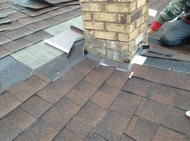 Roof with brown shingles, brick chimney, and metal flashing being installed. A person is kneeling.