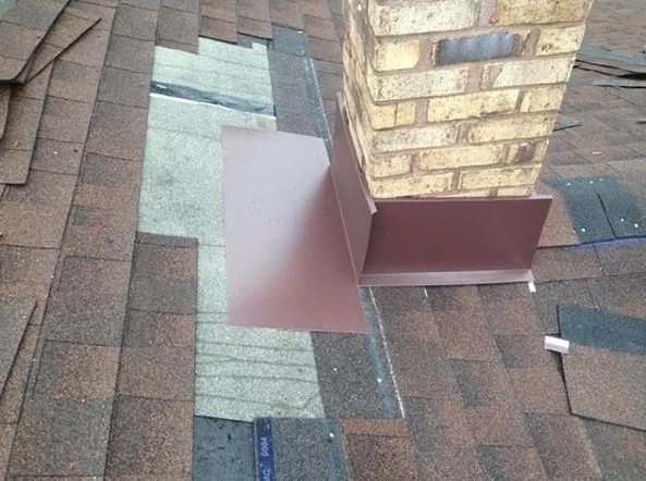 Chimney flashing on a shingled roof, showing brown metal against brown, gray, and tan shingles.