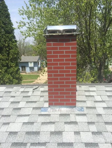 Brick chimney with a metal cap on a gray shingled roof, trees and a blue house in the background.