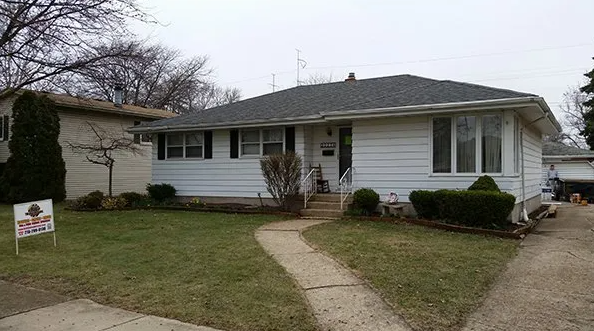 White ranch-style house with dark shutters, gray roof, and a front lawn. A for-sale sign is visible in the yard.