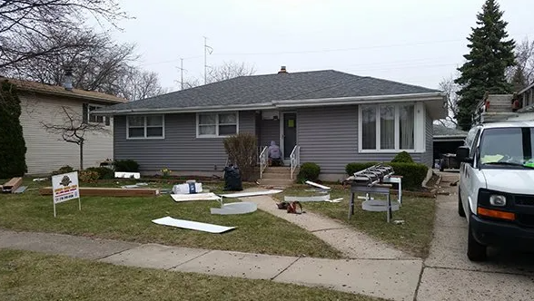 Gray house with construction materials in front yard; white van parked on right.