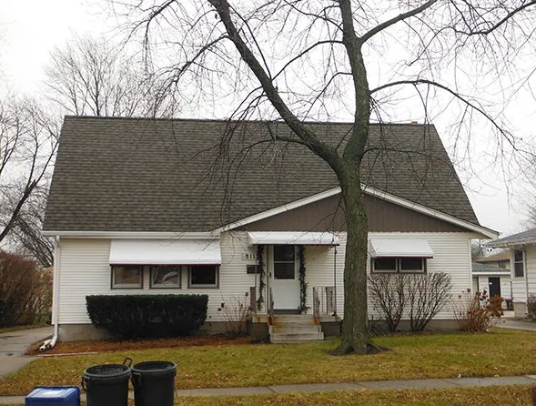 Small white house with brown roof and awning over the front door. Black trash cans in front.