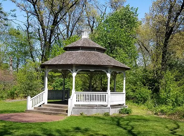 White gazebo with brown roof in a grassy park, surrounded by trees under a blue sky.