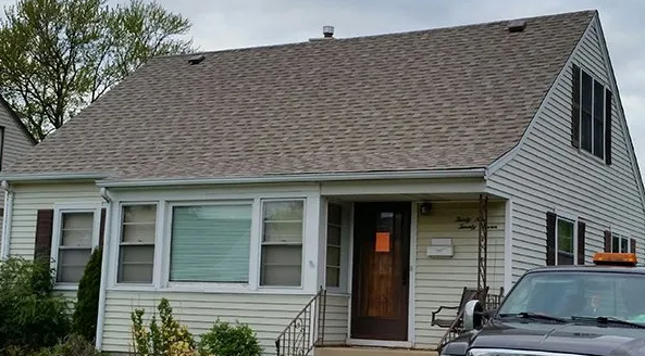 House with light siding, brown roof, windows, and a dark front door. A vehicle is parked in front.