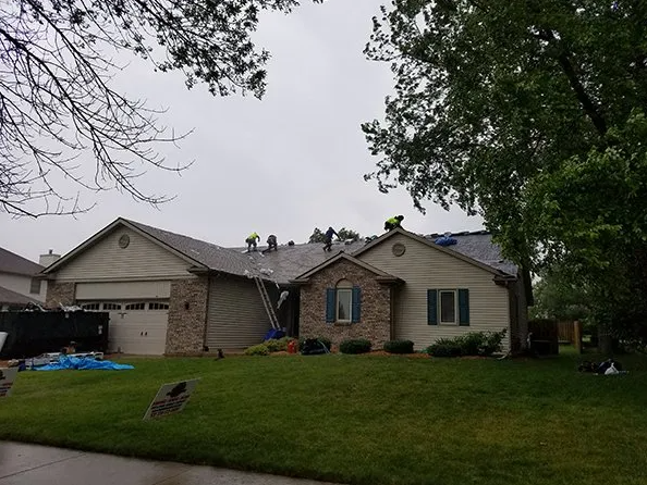 Roofers working on a residential roof, under a cloudy sky. House has brick facade and green lawn.