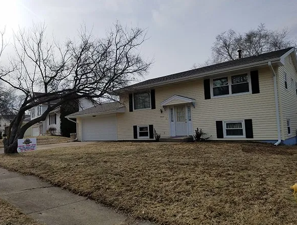 Beige two-story house with black shutters, attached garage, and bare tree in front yard.