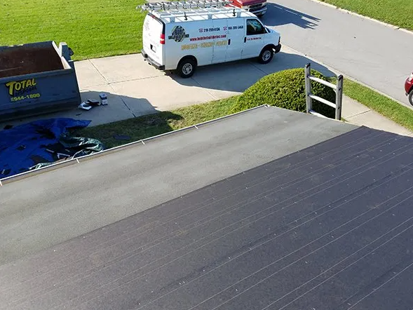 A black flat roof with a white van and a dumpster parked nearby on a sunny day.