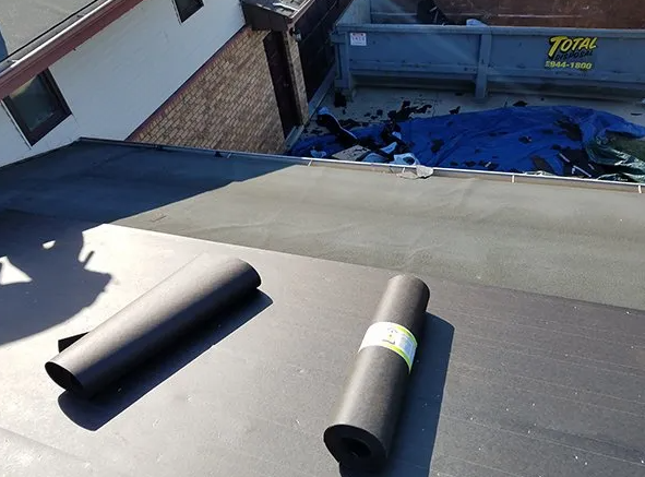 Two rolls of black roofing material on a flat roof, debris in background, next to a building.