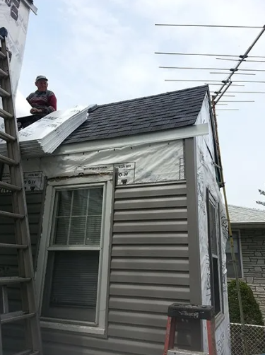 Man on roof installing shingles; gray siding and windows, outdoor setting.