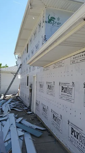 Building exterior under construction; plywood siding, white trim, and debris on the ground.