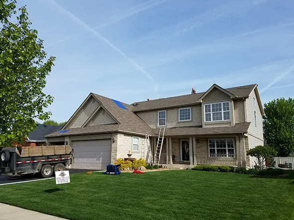 House with brown roof, beige siding, and green lawn. A trailer with building materials is parked on the left.