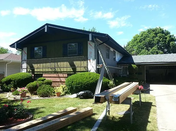 House exterior with ladder, lumber, and carport, under construction. Green bushes and blue sky.