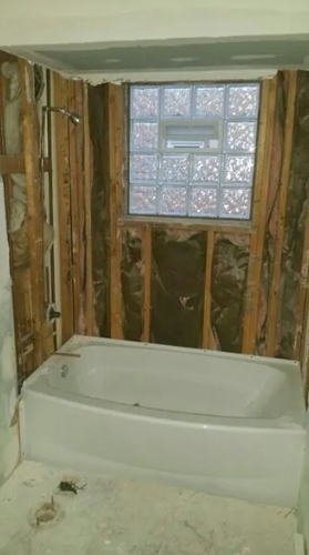 Bathroom under renovation: bathtub, shower fixture, glass block window, exposed wooden studs.