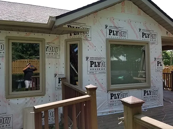 House exterior under construction, windows installed, wrapped in protective material; a person taking a photo is reflected in a window.