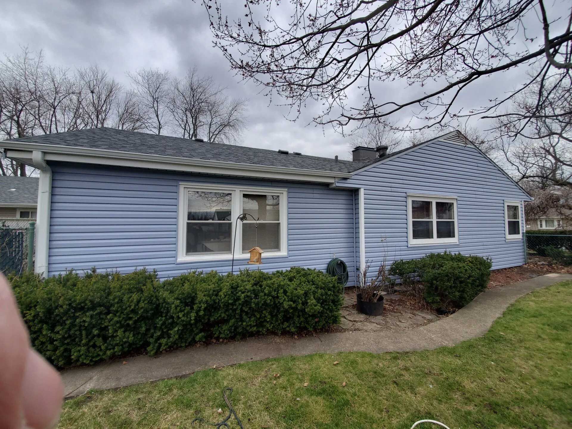 Blue-sided house with white-framed windows, surrounded by green shrubs and a lawn under an overcast sky.