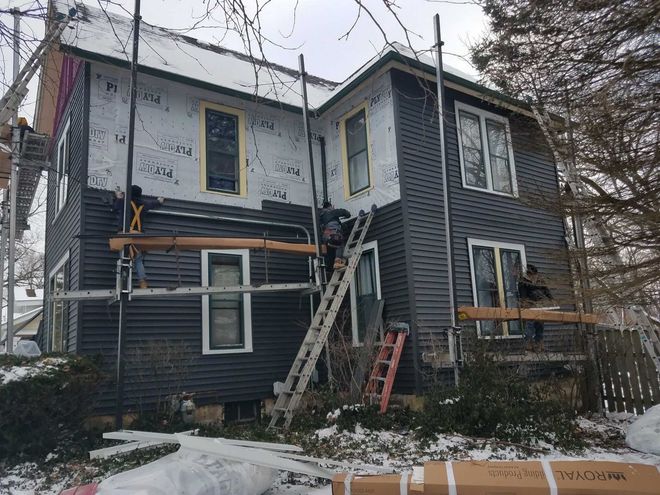 House exterior being painted, with scaffolding and workers. Dark siding, snow on ground, cloudy day.