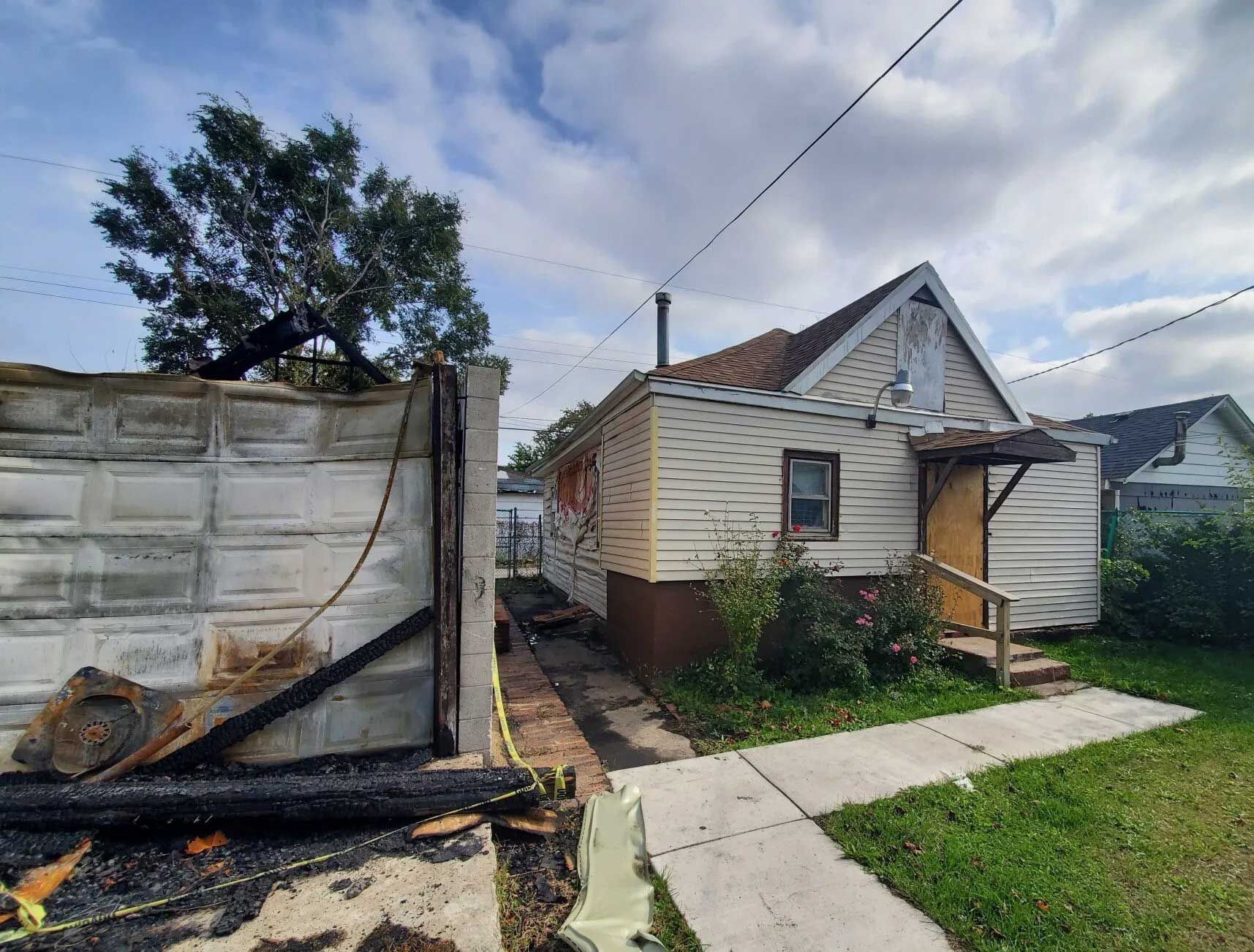 Small house with beige siding, a brown foundation, and a walkway next to a burned wall under a cloudy sky.