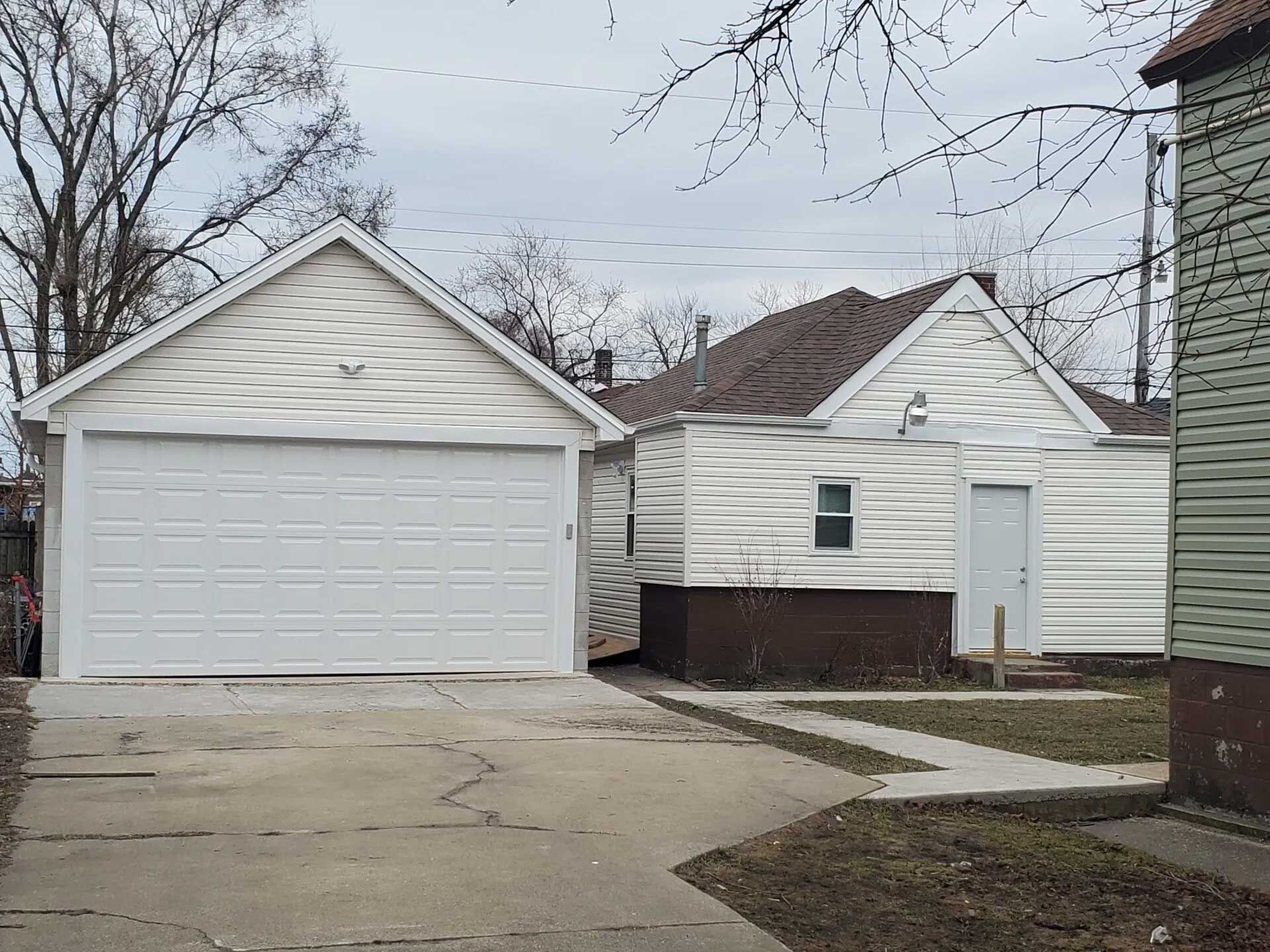 White garage with a sloped roof and driveway next to a smaller white building and a sidewalk.