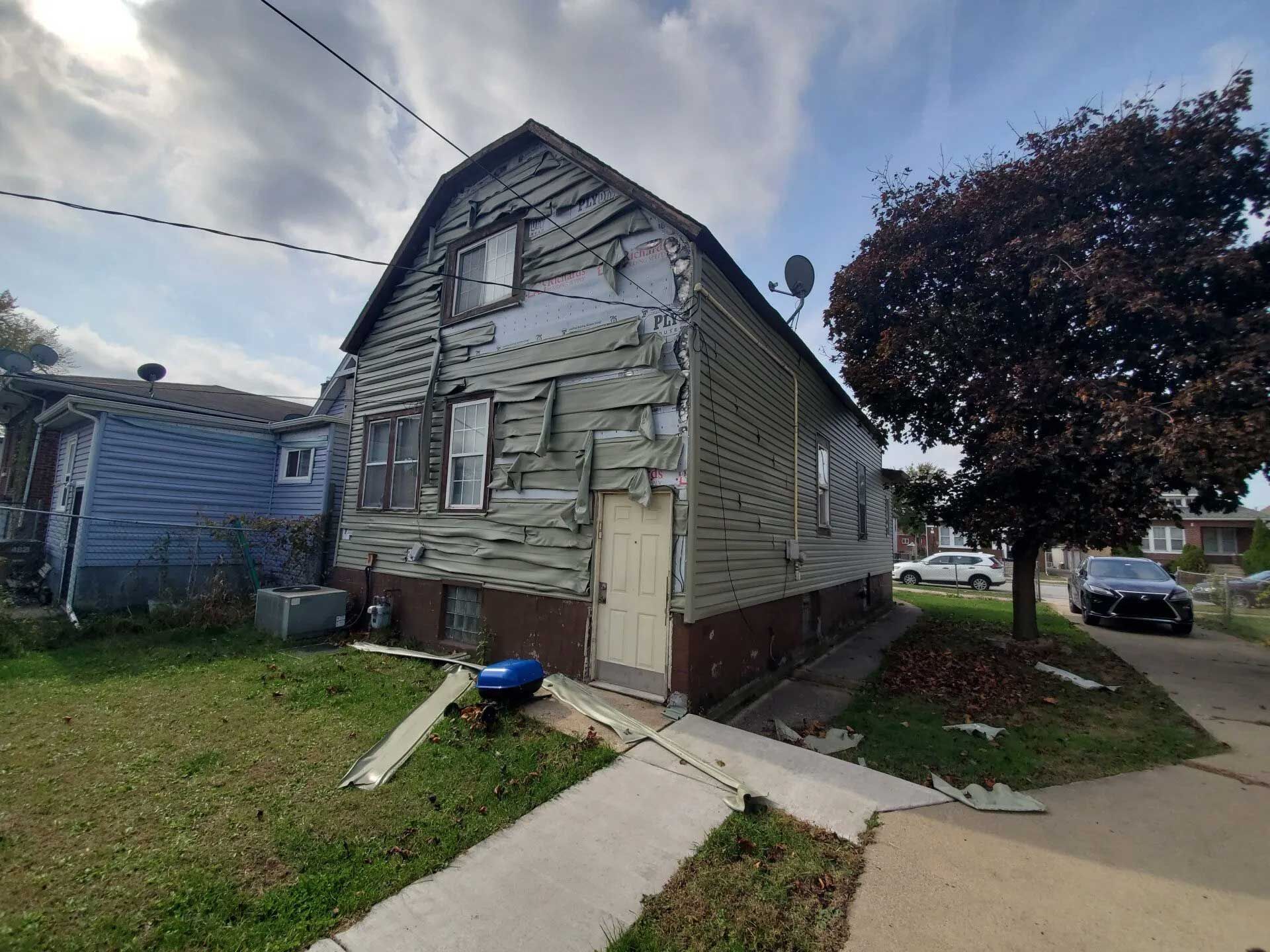 Dilapidated house with peeling siding, set on a grassy lot, sidewalk in front, overcast sky.