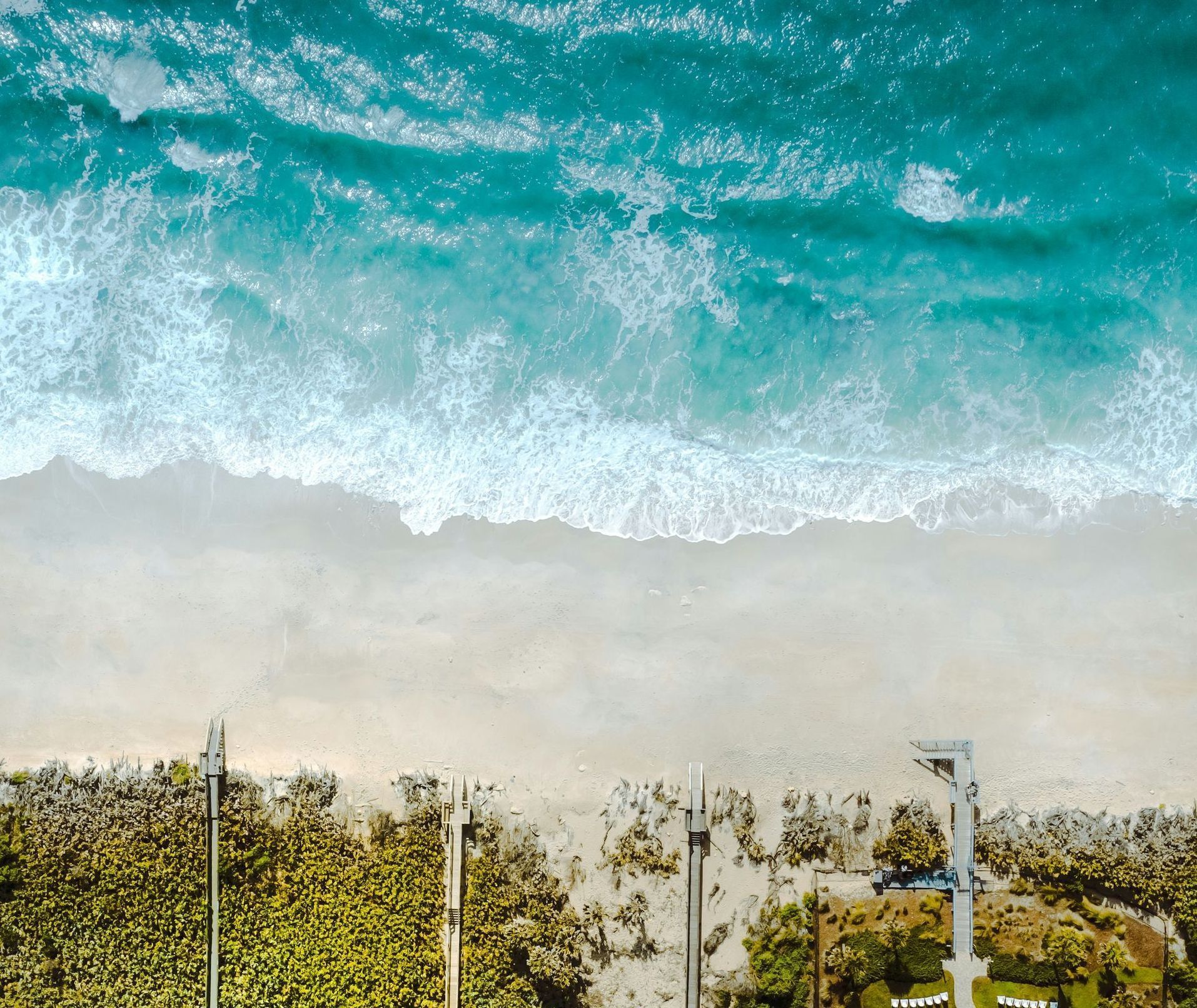 Aerial view of turquoise ocean waves above a sandy beach with rows of lounge chairs and umbrellas.
