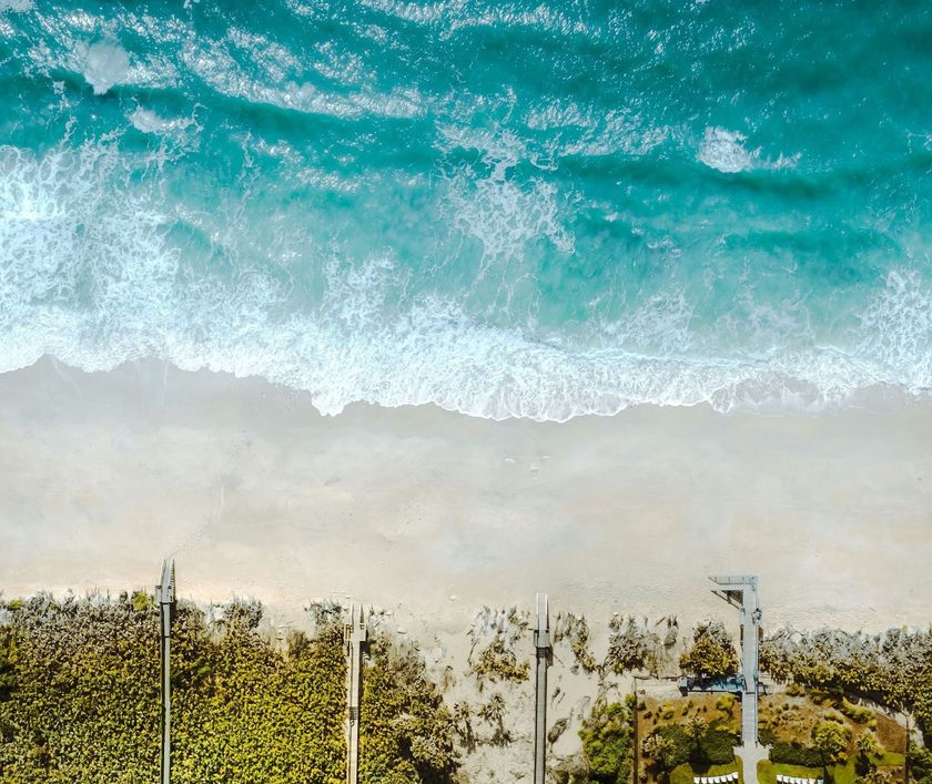 Aerial view of turquoise ocean waves above a sandy beach with rows of lounge chairs and umbrellas.