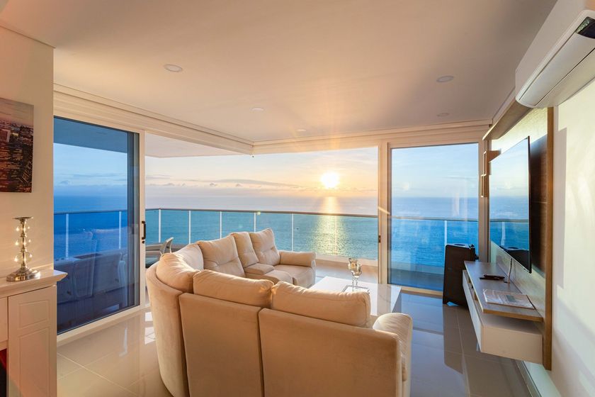 Modern living room with beige sofas overlooking an ocean sunset through floor-to-ceiling windows