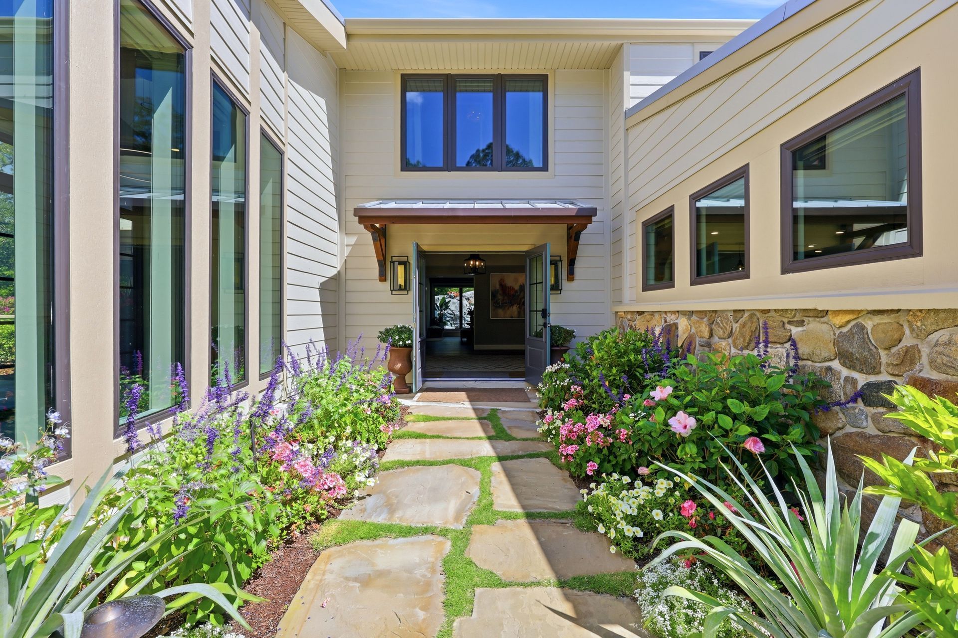 Front walkway to a modern house, with flower beds, stone path, and large windows.