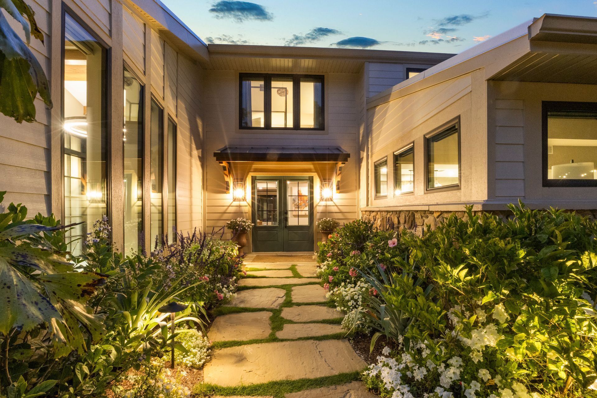 Modern two-story house at dusk with lit windows and a stone path through lush landscaping