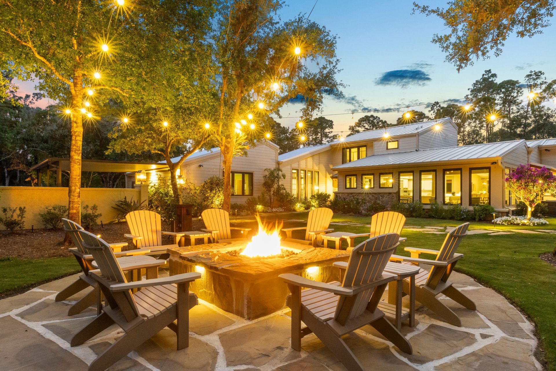 Backyard patio with fire pit, Adirondack chairs, string lights, and a lit house at dusk